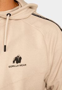 Lichtbeige hoodie met een gestructureerde stof. "Gorilla Wear" logo in het zwart op de borst en schouder, met contrasterende zwarte accenten.