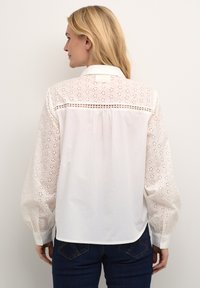 Bluză albă cu m�âneci lungi și bufante, cu broderie cu ochiuri. Guler și închidere cu nasturi, cu un tiv ușor curbat. Material ușor.
