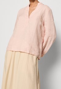 Lichtroze linnen blouse met lange mouwen en een v-hals, gecombineerd met een crèmekleurige geplooide rok. Zachte textuur en een ontspannen pasvorm.