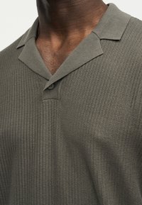 Πλεκτό πόλο σε σκούρο ελαιδί πράσινο με υφή, με ένα μόνο κουμπί στον γιακά, φορεμένο από ένα άτομο.