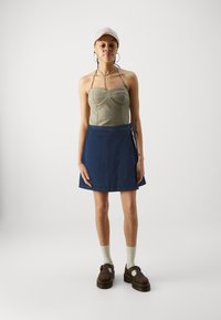 Olivgrön strapless topp, mörk denimomslagskjol, bruna platåskor och grå strumpor; modellen står mot en enfärgad bakgrund.
