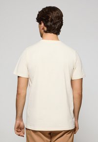 Camiseta corta de manga corta de color crema, hecha de algodón, con cuello redondo y un corte relajado. La vista desde atrás muestra una textura simple y suave.