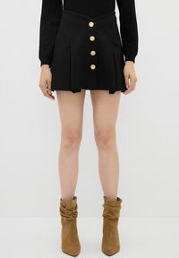 Jupe mini plissée noire avec cinq boutons dorés sur le devant, présentant une taille ajustée et une texture lisse. Associée à des bottines beiges.