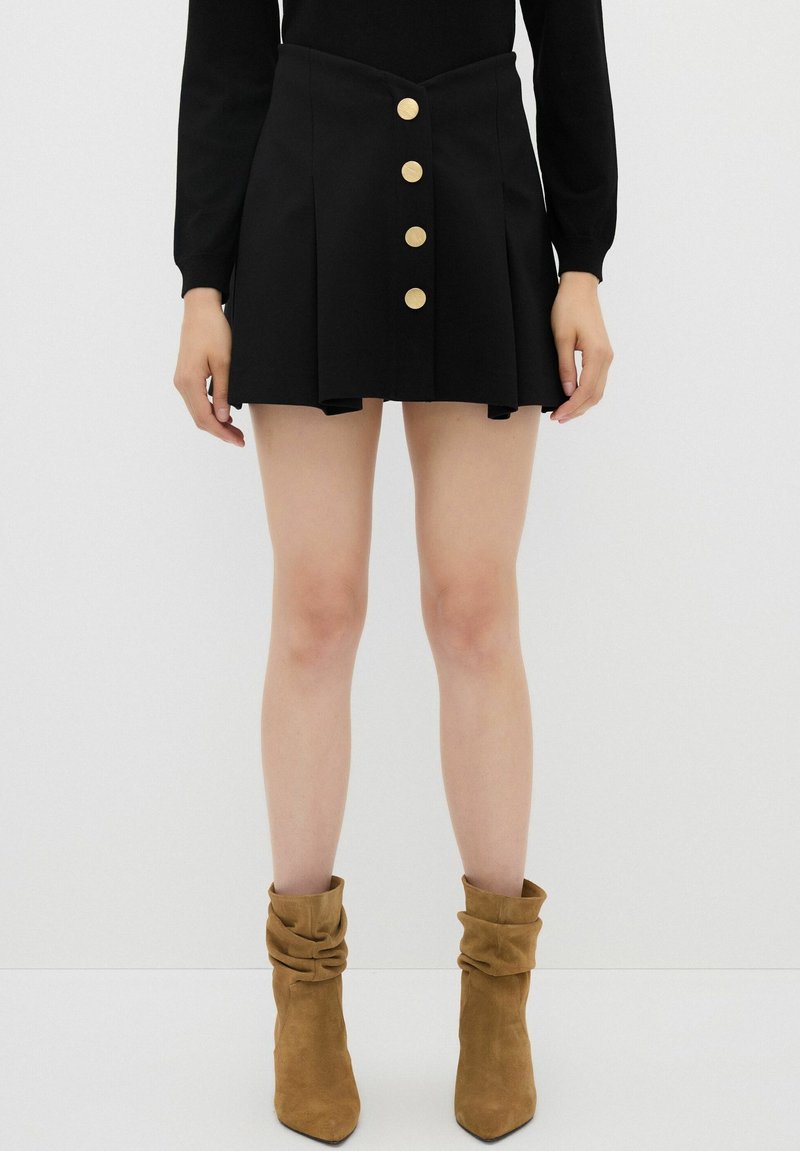 Jupe mini plissée noire avec cinq boutons dorés sur le devant, présentant une taille ajustée et une texture lisse. Associée à des bottines beiges.