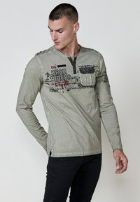 Olíva zöld, hosszú ujjú póló, henley nyakkivágással, grafikai nyomattal, zsebbel a mellkasán és kontrasztos varrási részletekkel.