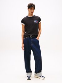 Svart T-skjorte med hvite rulleermene, med et fargerikt merke. Blå baggy jeans med oppbrettede kanter, sammen med beige sneakers.