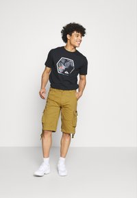 Svart T-skjorte med grafisk design, kombinert med brune cargo-shorts med sidelommer og justerbare hems, avsluttet med hvite sneakers.