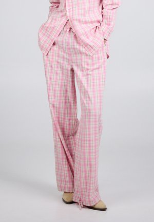 Person som har på seg løse rosa rutete bukser og matchende skjorte, står med hendene i lommene og beige sko synlige.