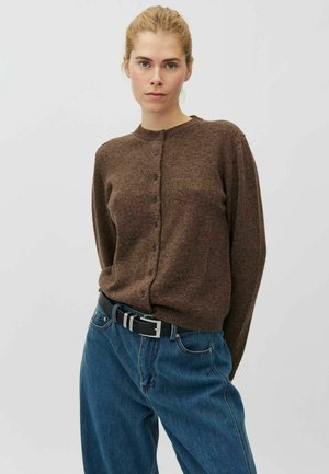 Femme aux cheveux blonds attachés, portant un pull marron boutonné et un jean bleu ample avec une ceinture noire sur un fond uni et clair.