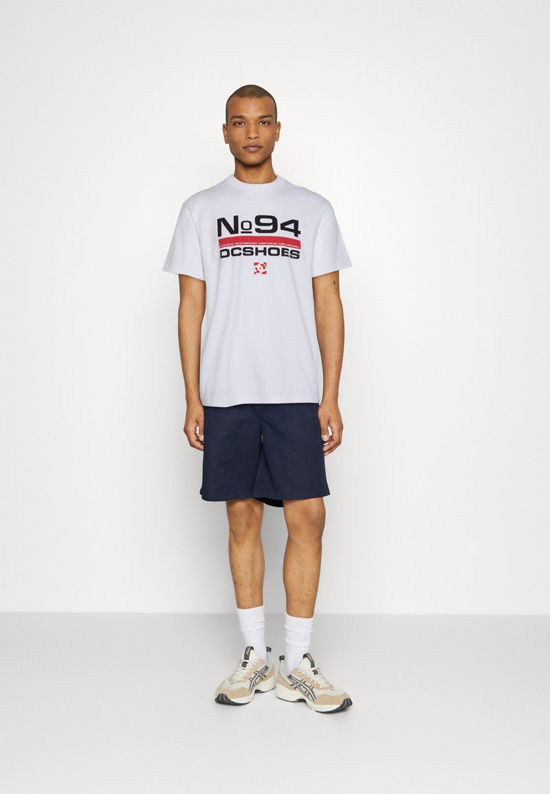 T-shirt bianca con testo grafico nero e rosso, abbinata a pantaloni corti blu navy. Il modello indossa sneakers beige e calzini bianchi. Design atletico semplice.