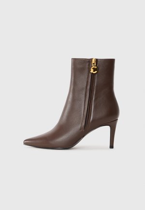 Bottine en cuir marron avec un bout pointu, un talon aiguille fin et une fermeture éclair sur le côté avec un accent doré. Texture lisse, design épuré.