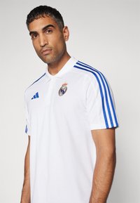 Maglietta polo bianca con righe blu sulle maniche, caratterizzata da un colletto e una pattina con bottoni. Logo del Royal Madrid sul petto. Tessuto dalla texture liscia.