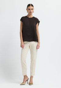 Top corto marrone a maniche corte con dettagli a bottone sulle spalle, abbinato a pantaloni crema su misura e scarpe beige con tacco alto su uno sfondo neutro.