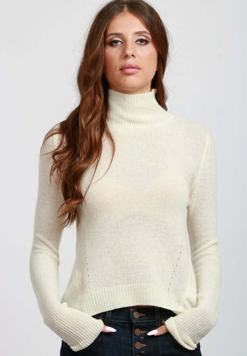 Donna con lunghi capelli castani che indossa un maglione a collo alto crema e jeans blu scuro con bottoni, in posa con le mani nelle tasche.