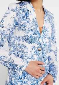 Blazer bianco con stampe botaniche e animali blu. Realizzato in un tessuto leggero, presenta una chiusura con un solo bottone e un collo a punta.