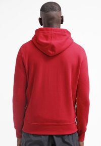 Röd huva tröja med en slät textur, som har en känguruficka, ribbade ärmar och nederkant, visad framifrån.