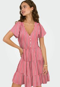 Femme debout, main sur la hanche, portant une robe à manches courtes à rayures verticales rouges et blanches avec un col en V, des boutons et des bijoux.