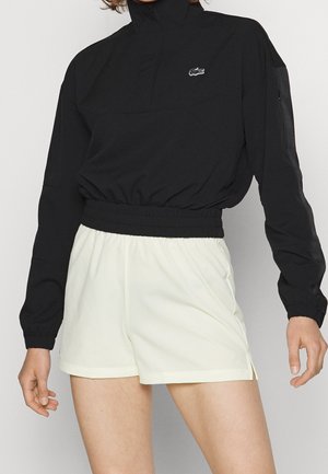 Personne portant un pull court noir à manches longues et un short blanc ample, debout devant un fond clair uni.