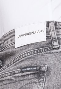 Jeans in denim grigio con un'etichetta in pelle bianca che riporta la scritta "CALVIN KLEIN JEANS." Presentano dettagli di cucitura e una piccola etichetta nera su una tasca.