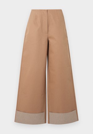 Nő barna ráncolt hosszú ujjú felsőt, bézs színű, széles szárú nadrágot kockás mandzsettával, fekete cipőt visel, és fekete szegecses keresztpántos táskát hord.