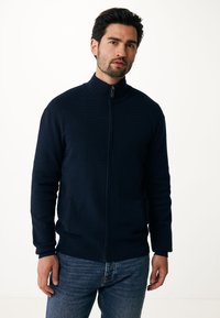 Světle modrý svetr na zip s texturovaným vzorem, vysokým límcem a dvěma předními kapsami, spárovaný s světle modrými džíny.
