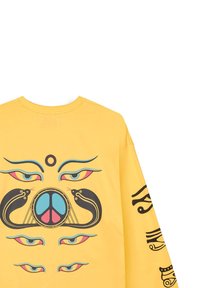 Camiseta de manga larga amarilla con gráficos intrincados en negro y colores, que incluyen ojos, serpientes y un símbolo de paz. Material suave de algodón.