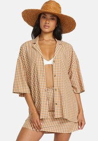 Brown and cream checkered oversized shirt with short sleeves, button front, paired with a matching drawstring mini skirt, and a wide-brimmed straw hat.