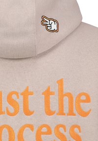 Beige hoodie featuring orange text "Just the Process" and a white, orange-accented peace sign embroidery on the hood. Soft texture, relaxed fit.