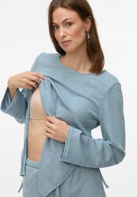 Top in tessuto azzurro chiaro, con texture e maniche lunghe; il modello mostra una sottoveste beige sottostante. Design avvolgente notevole con nastri.