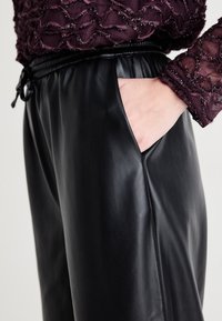 Pantalons en cuir synthétique noir avec une taille élastique et un cordon de serrage, accompagnés de poches latérales, associés à un haut texturé violet.
