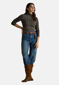 Kobieta stojąca z ręką na biodrze, ubrana w wzorzystą bluzkę z długim rękawem, niebieskie dżinsy z wysokim stanem, brązowy pasek i brązowe zamszowe botki.