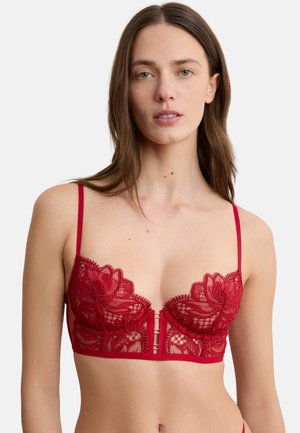 Donna che indossa un reggiseno rosso in pizzo con ricamo floreale e spalline sottili, su uno sfondo grigio chiaro uniforme.
