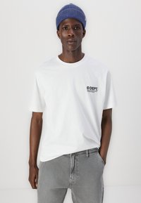 Λευκό βαμβακερό μπλουζάκι με κοντά μανίκια και στρογγυλή λαιμόκοψη, με μικρό μαύρο λογότυπο στο αριστερό στήθος. Υφασμάτινο μπλε πλεκτό καπέλο.