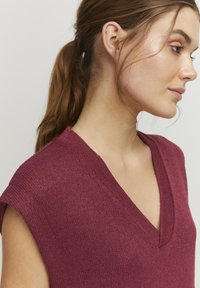 Haut en tricot bordeaux avec un décolleté en V profond et des manches courtes. Le tissu a une finition texturée, offrant une apparence légère et décontractée.