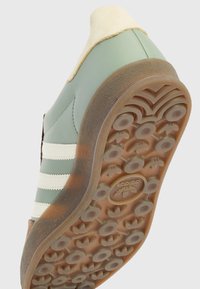 Zelené kožené tenisky s bílými pruhy, nubukovou patní částí a texturovanou gumovou podrážkou se zaoblenými vzory a logem uprostřed.