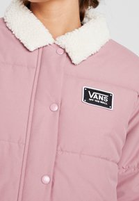 Lyserød pufferjakke med en kontrasterende hvid, struktureret pelskrave. Har trykknapper og et sort logo-patch med mærkenavnet.