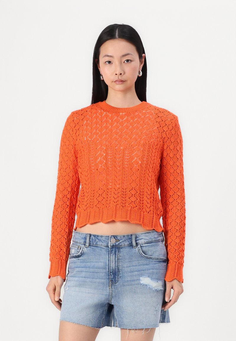 Maglione cropped arancione a maglia con un motivo a rombi e orlo a costine. Abbinato a pantaloni shorts in denim chiaro con una finitura distressed.