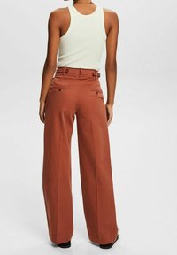Pantalon à jambes larges de couleur orange rouille, fabriqué dans un tissu lisse. Il présente une taille haute, des passants pour ceinture et deux poches arrière avec fermetures à boutons.