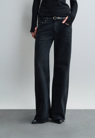 Person wearing high-waisted black wide-leg jeans with black belt and black pointed shoes, hand in pocket, standing against plain wall.
