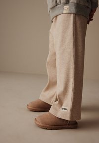 Béžové rebrované nohavice s voľným strihom a značkovou nášivkou, v kombinácii s hnedými semišovými slip-on topánkami. Mäkká textúra, viditeľné švy a široké nohavice.