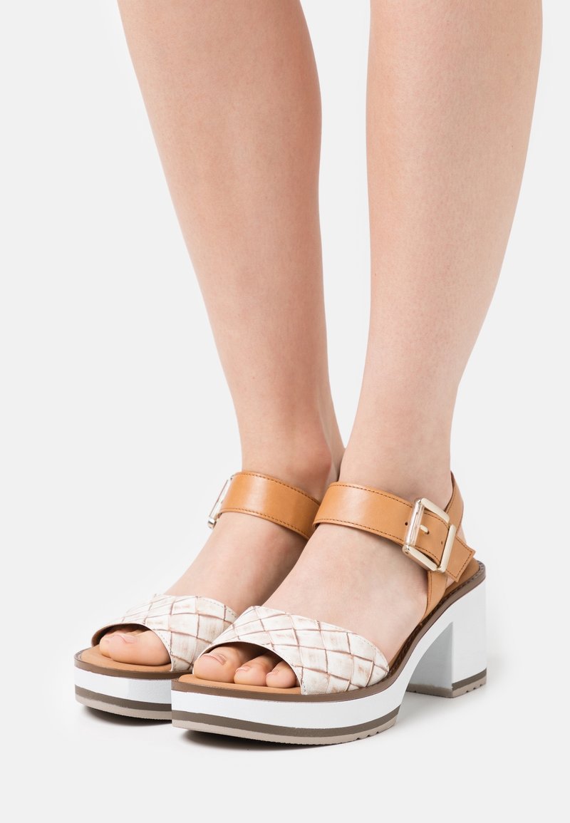 Pieds portant des sandales compensées beige et blanches avec des brides avant tressées et des brides de cheville avec boucles argentées sur fond blanc.