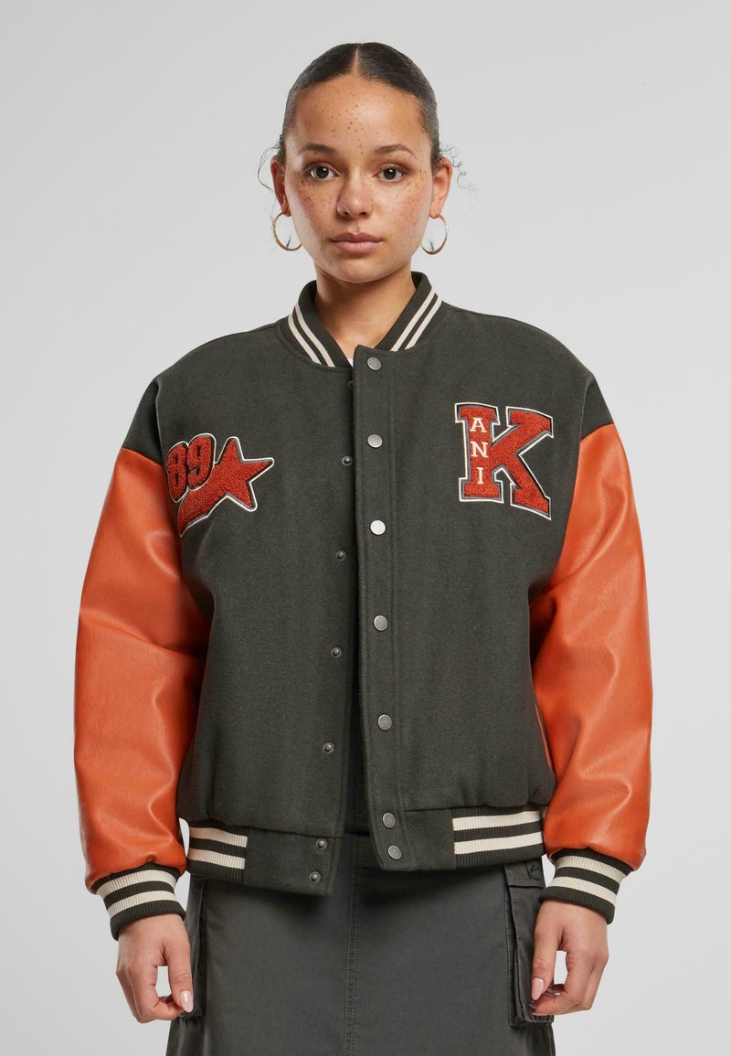 Varsityjakke i mørkegrønn og oransje, med trykknappfront, stripete mansjetter og broderte lapper, som viser en lagdelt tekstur.