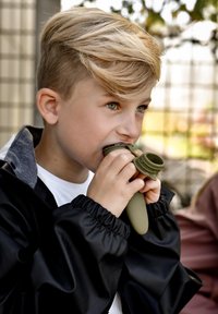 Otrok drži olivno zeleno silikonsko igračo ob ustih, oblečen v črno jakno z nabranimi manšetami in sivim ovratnikom, z kratkimi svetlimi lasmi.