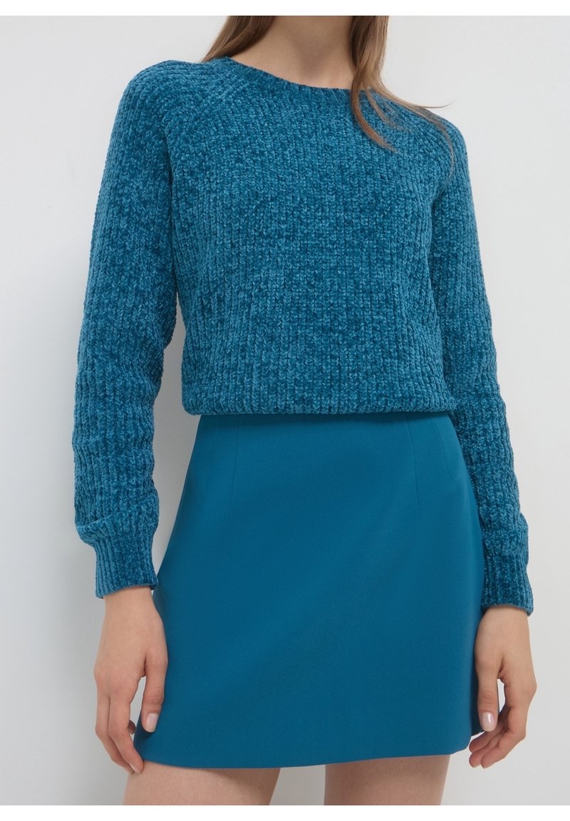 Femme portant un pull en maille bleu texturé et une mini-jupe unie bleue assortie, debout devant un fond clair uni.