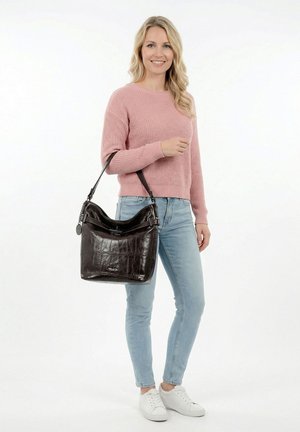 Nainen vaaleanpunaisessa paidassa ja vaaleissa farkuissa kantaa suurta mustaa nahkaista olkalaukkua, hymyilee ja seisoo valkoista taustaa vasten.