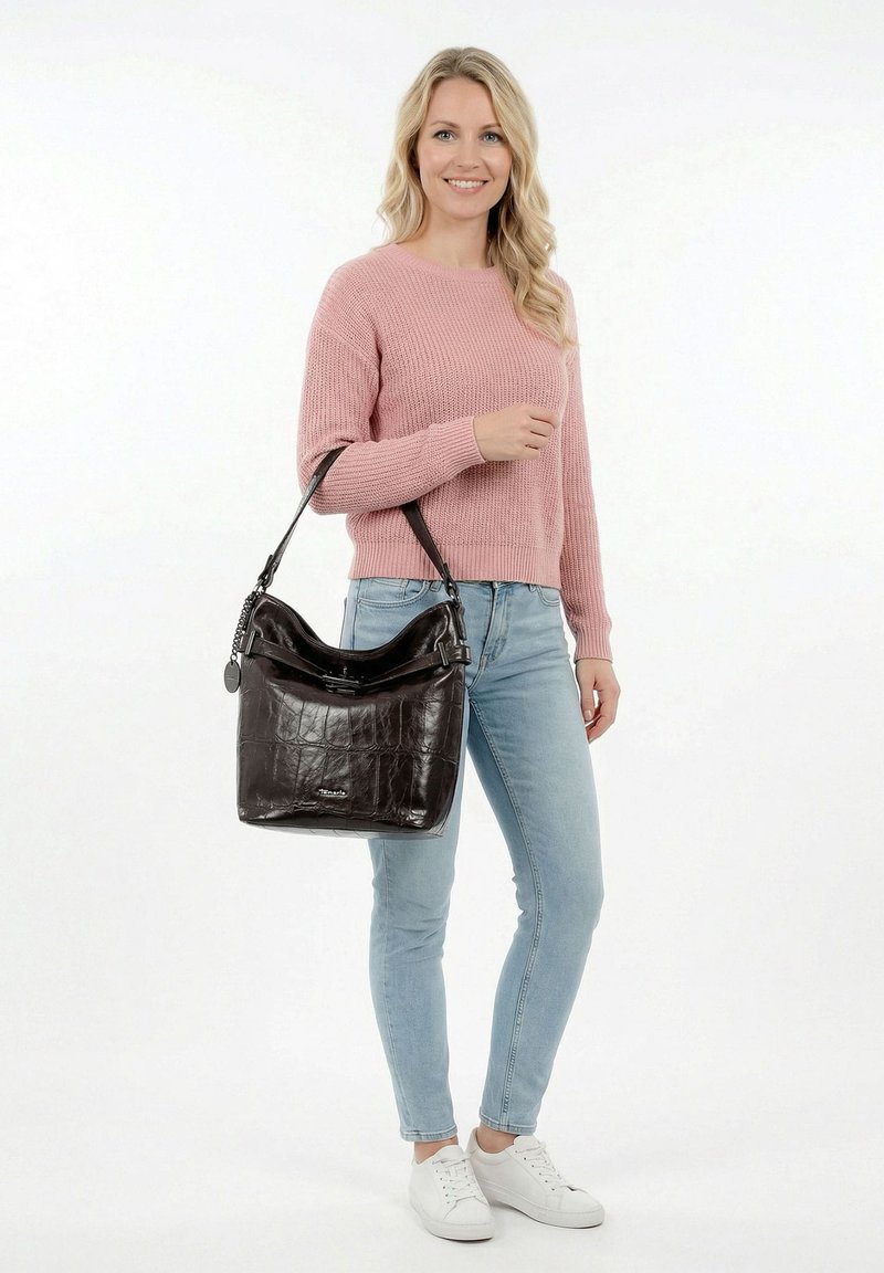 Žena v růžovém svetr a světlých džínách nesoucí velkou černou koženou kabelku přes rameno, usmívající se a stojící před bílým pozadím.