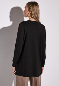 Svart långärmad topp med rundad halsringning, sidslitsar och mjuk textur, bärs med ljusbeiga byxor. Bakifrån fångat.