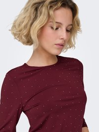Haut à manches longues bordeaux en tissu doux, présentant un motif à pois avec de petits accents blancs, col rond et coupe ajustée.