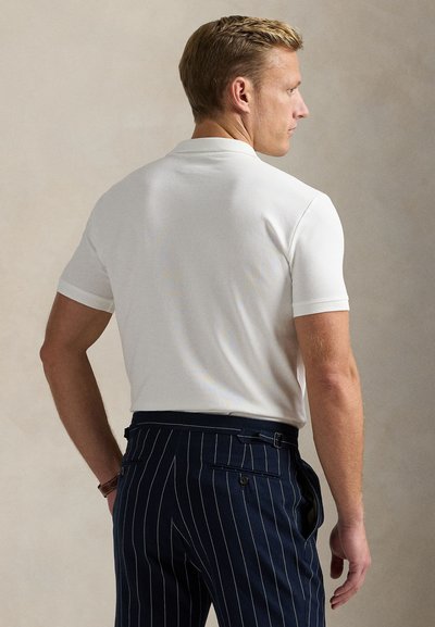 Baltas trumpomis rankovėmis polo marškinėlis su lygiu audiniu, dėvimas su tamsiai mėlynais dryžuotais kelnėmis. Turi klasikinius apykaklę ir subtilius briaunuotus rankogalius.