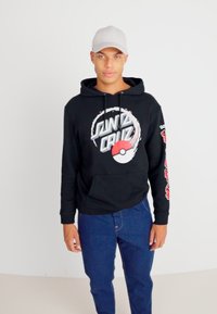 Black cotton hoodie with "Santa Cruz" and Pokéball graphic. Features a front pocket, drawstrings, and graphic accents on the sleeves. Blue jeans.